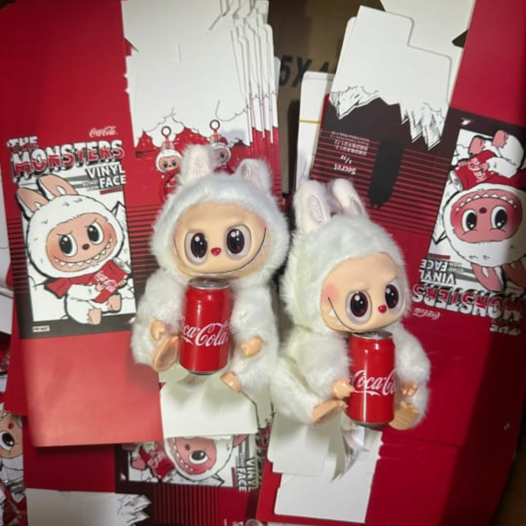 Two dolls in white outfits holding Coca-Cola cans with packaging in the background.