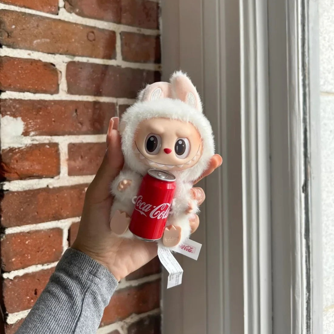 Plush toy resembling a character holding a Coca-Cola can against a brick wall.
